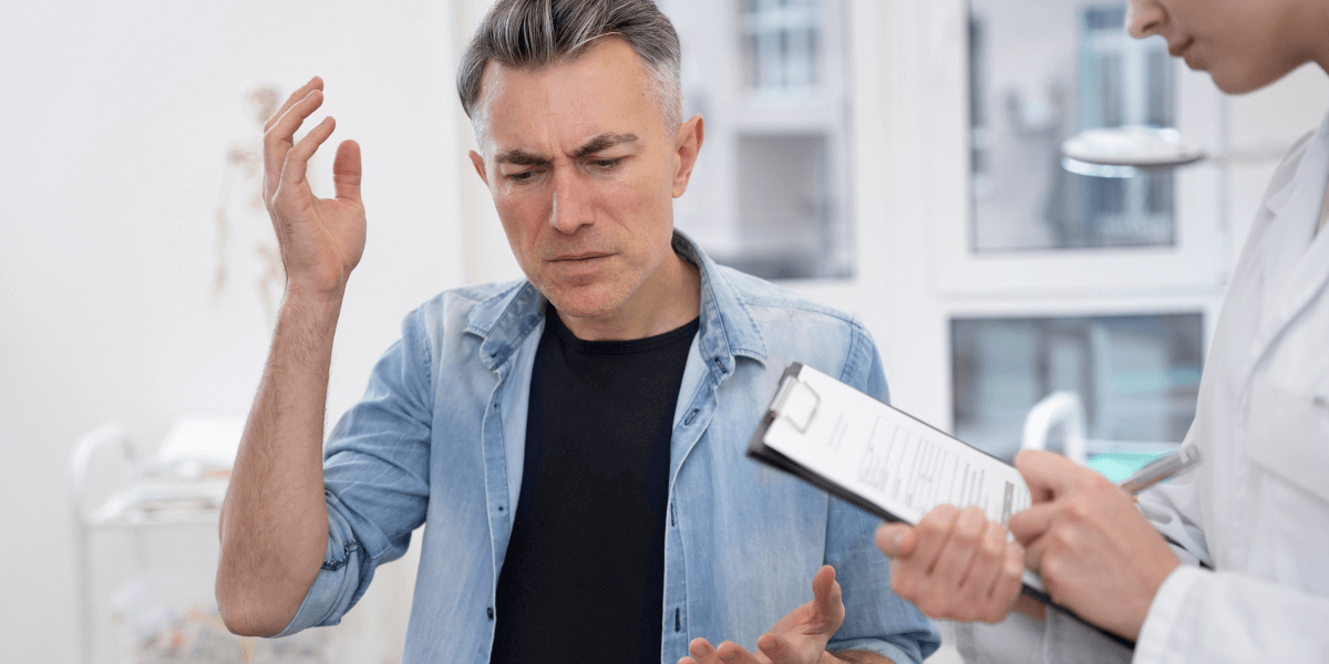Middle-aged man looking unwell and touching his head while consulting a doctor who is holding a clipboard."